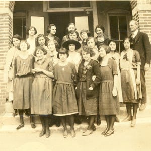 May include: A sepia-toned vintage photograph depicting a large group of individuals standing in front of a brick building. The group consists of many people wearing dresses with drop waists and one person in a suit, all posed for a formal portrait.