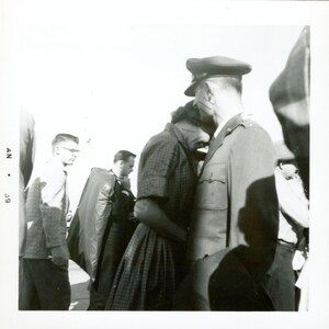 May include: A monochrome photograph shows a woman in a patterned dress embracing a man in a military uniform. Other people are visible in the background, suggesting a public location. The image has a vintage feel, evoking a sense of nostalgia and a bygone era.