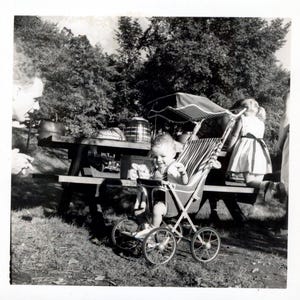 May include: A monochrome photograph showing a child seated in a vintage stroller with a striped canopy. The stroller has spoked wheels and is on grass near a picnic table. Another child is kneeling close by.