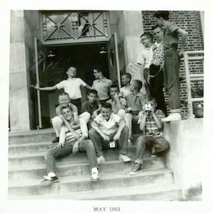 May include: Black and white photograph of a group of boys posing on steps in front of a building. Some are sitting, some are standing, and one is holding a camera. The text "MAY 1962" is at the bottom.