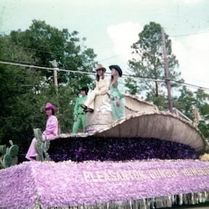 May include: A parade float shaped like a large cowboy hat, decorated with purple and lavender flowers. Several people in cowboy hats and colorful outfits are on the float. The text "Pleasanton Cowboy Homecoming" is visible on the float.