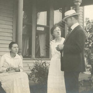 May include: Vintage black and white photograph of three people on a porch. A woman sits on the left, another woman stands in the middle, and a man in a suit and hat stands on the right. The porch has a wooden railing and a window.