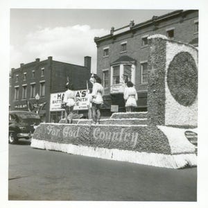 May include: A monochrome image of a parade float adorned with flowers, bearing the inscription "For God and Country". Several individuals in white attire are positioned on the float. Buildings and a vintage car are visible in the background, creating a historical scene.