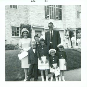 May include: Black and white photograph of a family posing in front of a building. The image shows a woman in a dress, a man in a suit, and several children in formal attire. The text "DEC 64" is in the top left corner.
