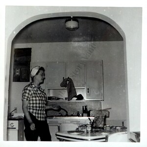 Könnte beinhalten: Schwarz-Weiß-Fotografie einer Vintage-Küchenszene. Eine Frau in einem karierten Hemd und Kopftuch steht in der Nähe eines Retro-Tisches. Schränke, ein Kühlschrank und eine Leuchte sind sichtbar und erinnern an eine Ästhetik aus der Mitte des Jahrhunderts.
