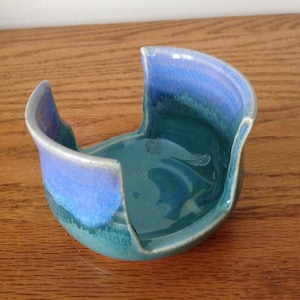May include: A handmade ceramic bowl with a unique, curved design. The bowl features a gradient of blue and teal glazes, creating a visually appealing piece. The bowl is approximately 6 inches wide and 4 inches tall.