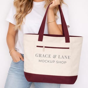May include: Cream-colored tote bag with burgundy accents and handles. The bag features a front pocket with a zipper and the text "GRACE & LANE MOCKUP SHOP" printed in black. The bag is held by a person.