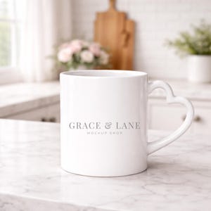 Op de afbeelding: Witte keramische mok met een hartvormig handvat. De mok heeft de tekst "GRACE & LANE MOCKUP SHOP" in grijs. De mok staat op een marmeren oppervlak.