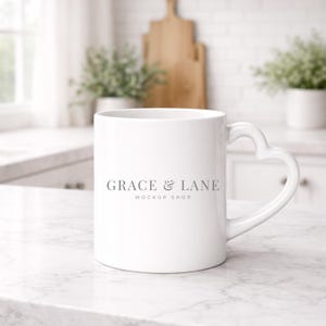 Op de afbeelding: Een witte keramische mok met een hartvormig oor. De mok heeft de tekst "GRACE & LANE MOCKUP SHOP" in grijs. De mok staat op een wit marmeren oppervlak, met een vage keukenachtergrond.