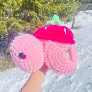 Könnte beinhalten: Eine handgefertigte rosa Häkelschildkröte mit einem leuchtend roten Erdbeerschild und grünen Blättern. Die Schildkröte hat ein schwarzes Auge. Die Schildkröte wird vor einem schneebedeckten Hintergrund gehalten.