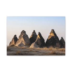 May include: A canvas print featuring a landscape of unique rock formations. The cone-shaped rocks are in varying shades of gray and brown, with some illuminated by sunlight. The sky is a gradient of light blue and yellow.