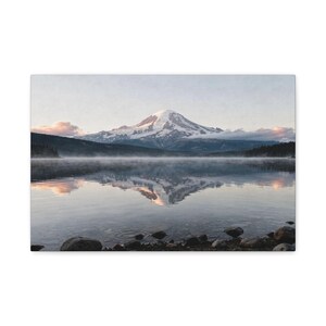 May include: A landscape canvas print featuring a snow-capped mountain reflected in a calm lake. The image captures a serene scene with a misty atmosphere, rocks in the foreground, and trees along the shoreline. The sky is a soft blue with hints of pink.