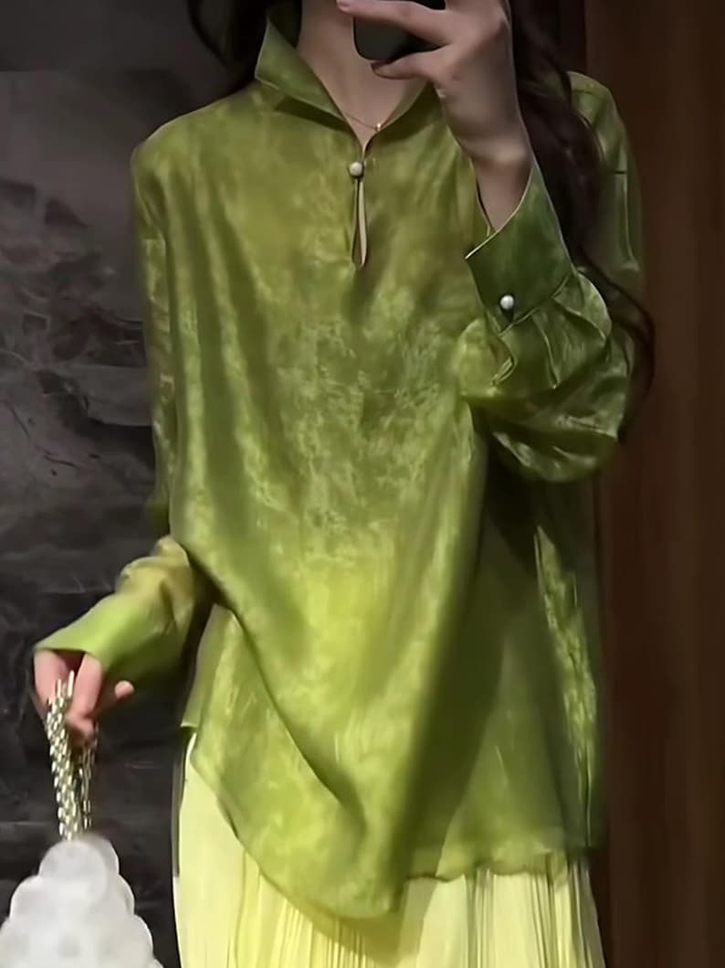 May include: A lime green blouse with a mandarin collar and long sleeves. The blouse has a keyhole neckline with a pearl button closure. The cuffs also feature pearl buttons. The blouse is paired with a light yellow pleated skirt.