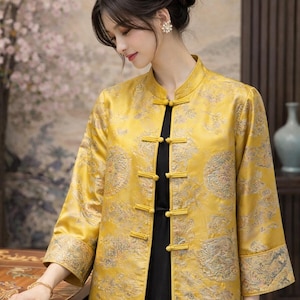 Puede incluir: Chaqueta amarilla de manga larga con cuello mandarín y cierres de rana. La chaqueta presenta un diseño ornamental y se lleva sobre un vestido negro. Las mangas son de tres cuartos.