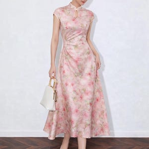 以下が含まれることがあります： ピンクの花柄ミディ丈ワンピース、マンダリンカラーとキャップスリーブ。フィットした胴体と流れるようなスカートが特徴です。ゴールドのハンドルが付いた白いハンドバッグとクリアヒールがルックを完成させます。