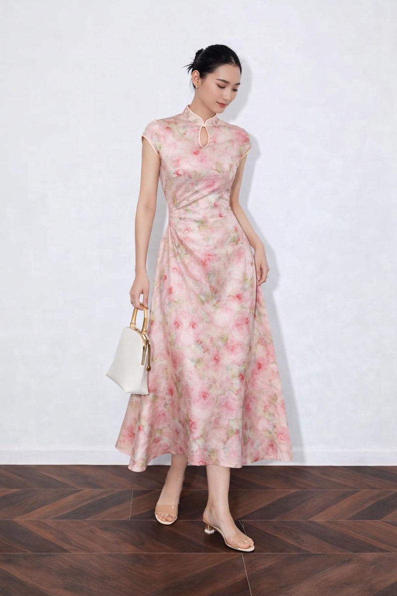 以下が含まれることがあります： ピンクの花柄ミディ丈ワンピース、マンダリンカラーとキャップスリーブ。フィットした胴体と流れるようなスカートが特徴です。ゴールドのハンドルが付いた白いハンドバッグとクリアヒールがルックを完成させます。