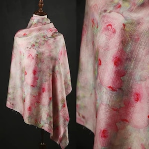 以下が含まれることがあります： ピンク色の花柄のスカーフ。マネキンにドレープされており、デザインが際立っています。生地は、ピンクと赤のバラと緑の葉の、水彩画のようなプリントが施されており、柔らかくロマンチックな美しさを作り出しています。