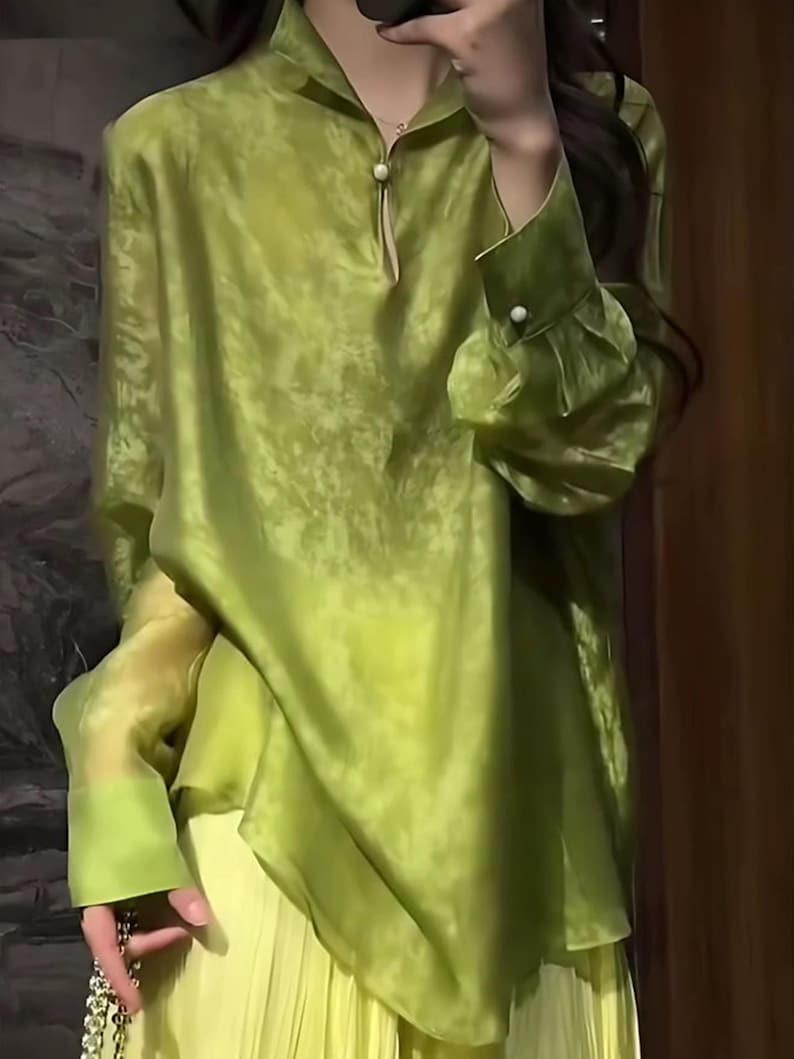 May include: A long-sleeved, lime green blouse with a mandarin collar and a small pearl button closure. The blouse has a flowing, semi-sheer fabric and is paired with a pleated, light yellow skirt. The sleeves have pearl button cuffs.