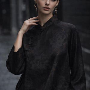 K&ouml;nnte beinhalten: Schwarzes Oberteil mit Stehkragen und floralem Muster. Das lang&auml;rmlige Oberteil hat einen Knopfverschluss. Die Person tr&auml;gt goldene Ohrringe und einen Ring.