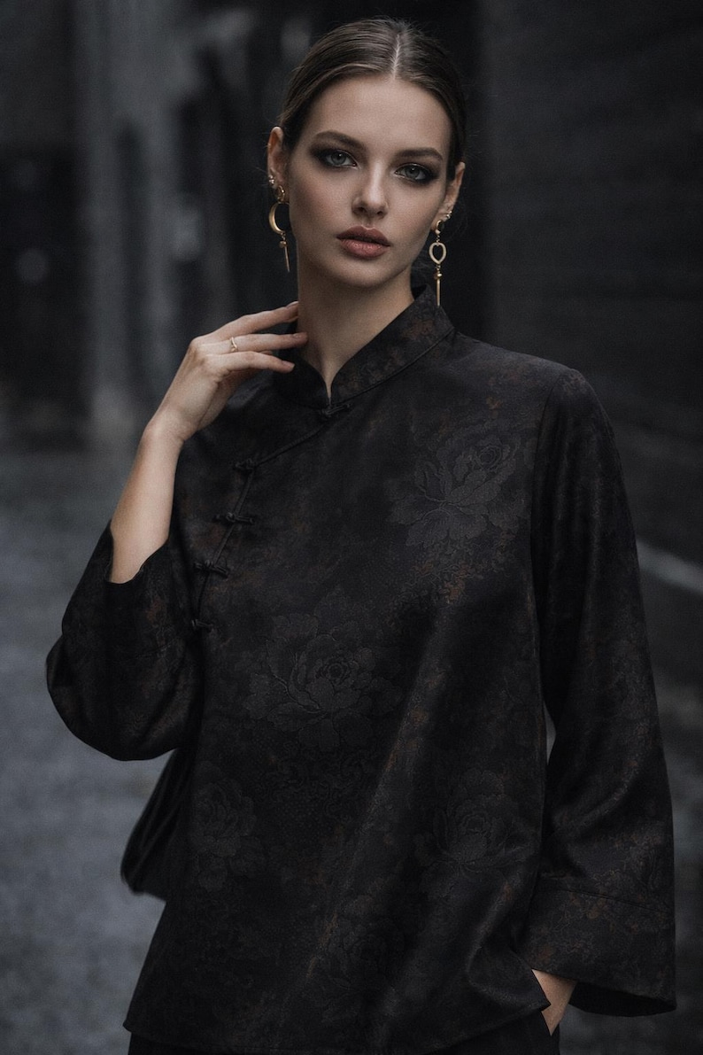 K&ouml;nnte beinhalten: Schwarzes Oberteil mit Stehkragen und floralem Muster. Das lang&auml;rmlige Oberteil hat einen Knopfverschluss. Die Person tr&auml;gt goldene Ohrringe und einen Ring.