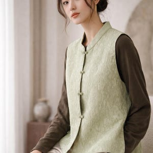May include: A light green vest with a textured pattern and a mandarin collar. The vest has four fabric-covered buttons down the front. The vest is worn over a long-sleeved brown shirt and paired with light-colored pants.