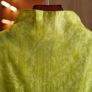 May include: Close-up of a lime green garment with a mandarin collar. The fabric has a subtle sheen and a textured appearance. The garment is hanging on a wooden hanger, suggesting it is a shirt or blouse.