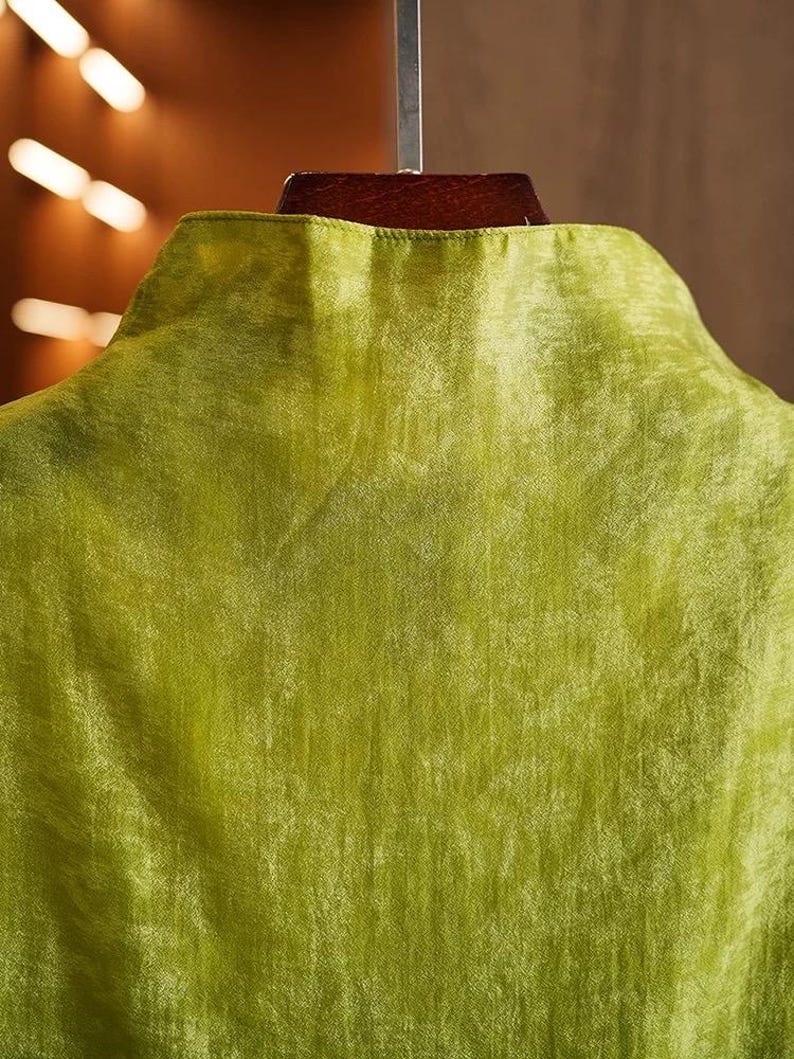 May include: Close-up of a lime green garment with a mandarin collar. The fabric has a subtle sheen and a textured appearance. The garment is hanging on a wooden hanger, suggesting it is a shirt or blouse.