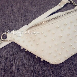 K&ouml;nnte beinhalten: Wei&szlig;e Bauchtasche mit silbernem Rei&szlig;verschluss und einer Kettenkette. Die Tasche ist mit kleinen wei&szlig;en Perlen bedeckt.