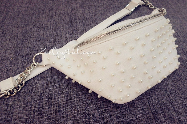 K&ouml;nnte beinhalten: Wei&szlig;e Bauchtasche mit silbernem Rei&szlig;verschluss und einer Kettenkette. Die Tasche ist mit kleinen wei&szlig;en Perlen bedeckt.