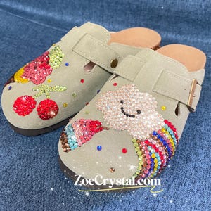 May include: A pair of beige suede clogs decorated with colorful rhinestones. One clog features a cherry design, while the other has a cloud with a smiley face and a rainbow. The clogs are embellished with sparkling rhinestones and have a website address, "ZoeCrystal.com", printed on the bottom.