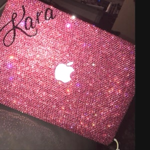 May include: A pink laptop covered in sparkling rhinestones. The Apple logo is visible in white. The name "Kara" is written in cursive script on the upper left corner. The overall effect is glamorous and eye-catching.