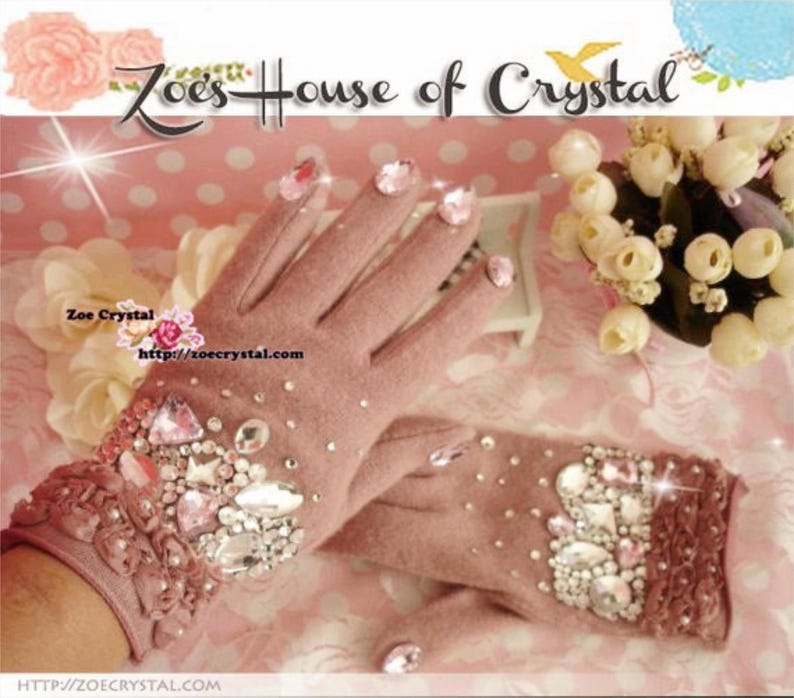May include: A pair of pink fingerless gloves with a lace cuff and a large cluster of rhinestones on the back of the hand. The gloves are decorated with pink and clear rhinestones. The text "Zoe's House of Crystal" is visible in the background.