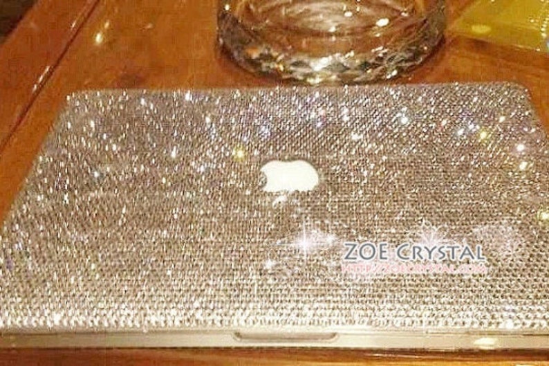 May include: A silver laptop computer covered in sparkling rhinestones. The laptop has a white Apple logo and the text "ZOE CRYSTAL" is visible on the bottom right corner.