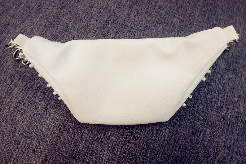 K&ouml;nnte beinhalten: Wei&szlig;e G&uuml;rteltasche aus Kunstleder mit einer silbernen Kette und Perlenbesatz.