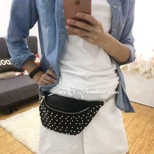 K&ouml;nnte beinhalten: Schwarze Bauchtasche mit wei&szlig;en Nieten, silbernem Rei&szlig;verschluss und Kette. Die Person tr&auml;gt ein wei&szlig;es T-Shirt, ein Jeanshemd und wei&szlig;e Shorts. Die Website 'ZoeCrystal.com' ist sichtbar.