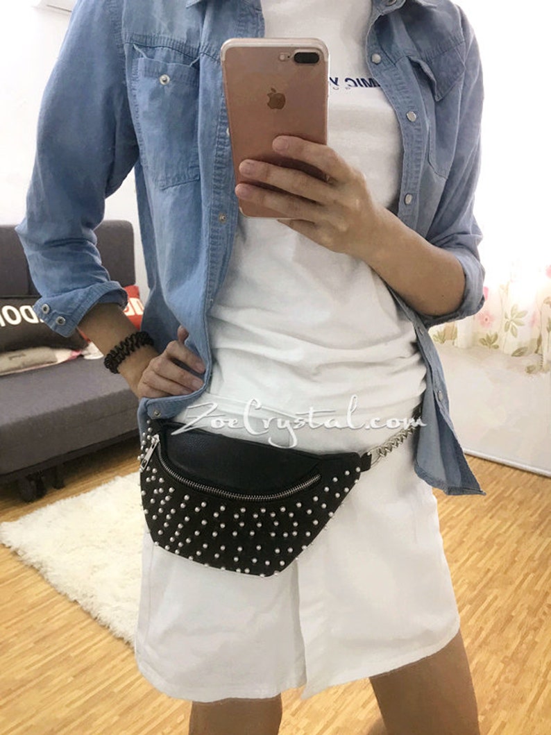K&ouml;nnte beinhalten: Schwarze Bauchtasche mit wei&szlig;en Nieten, silbernem Rei&szlig;verschluss und Kette. Die Person tr&auml;gt ein wei&szlig;es T-Shirt, ein Jeanshemd und wei&szlig;e Shorts. Die Website 'ZoeCrystal.com' ist sichtbar.