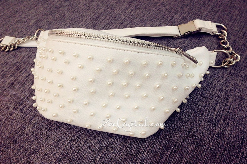 K&ouml;nnte beinhalten: Wei&szlig;e G&uuml;rteltasche aus Kunstleder mit silbernem Rei&szlig;verschluss und einer Kettenkette. Die Vorderseite der Tasche ist mit wei&szlig;en perlenartigen Perlen bedeckt.