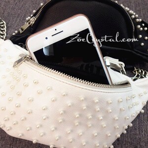 K&ouml;nnte beinhalten: Eine wei&szlig;e Bauchtasche mit Rei&szlig;verschluss und einem Kettenriemen. Die Bauchtasche ist mit kleinen wei&szlig;en Perlen besetzt. Im Hintergrund ist eine schwarze Lederbauchtasche mit Perlenbesatz zu sehen. Ein Smartphone befindet sich in der wei&szlig;en Bauchtasche.