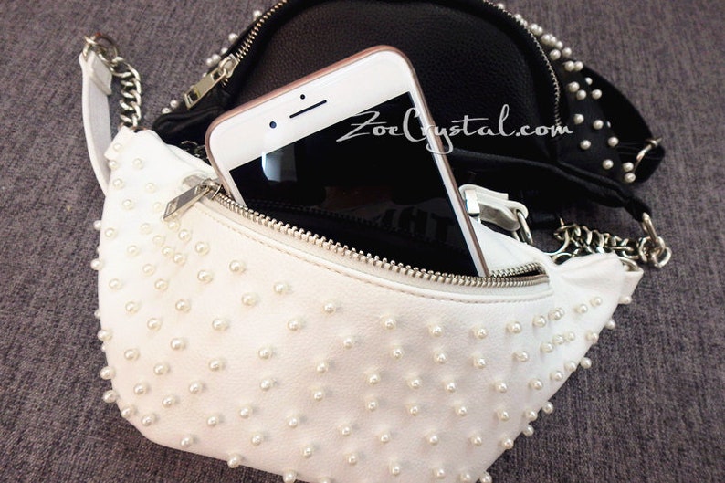 K&ouml;nnte beinhalten: Eine wei&szlig;e Bauchtasche mit Rei&szlig;verschluss und einem Kettenriemen. Die Bauchtasche ist mit kleinen wei&szlig;en Perlen besetzt. Im Hintergrund ist eine schwarze Lederbauchtasche mit Perlenbesatz zu sehen. Ein Smartphone befindet sich in der wei&szlig;en Bauchtasche.