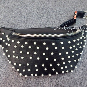 K&ouml;nnte beinhalten: Schwarze Bauchtasche mit wei&szlig;en, perlenartigen Nieten. Die Tasche hat einen silbernen Rei&szlig;verschluss und einen silbernen Kettenriemen. Der verstellbare Riemen ist schwarz. Der Text "ZoeCrystal.com" ist sichtbar.