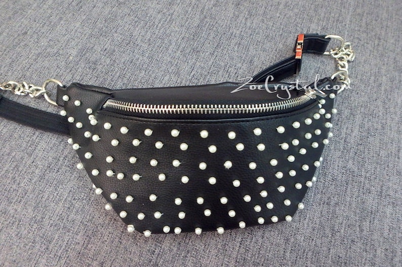 K&ouml;nnte beinhalten: Schwarze Bauchtasche mit wei&szlig;en, perlenartigen Nieten. Die Tasche hat einen silbernen Rei&szlig;verschluss und einen silbernen Kettenriemen. Der verstellbare Riemen ist schwarz. Der Text "ZoeCrystal.com" ist sichtbar.
