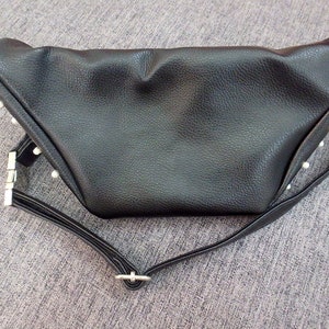 K&ouml;nnte beinhalten: Schwarze Bauchtasche mit strukturierter Oberfl&auml;che und dreieckiger Form. Die Tasche hat einen langen, verstellbaren Riemen und silberfarbene Beschl&auml;ge. Kleine, wei&szlig;e, perlenartige Verzierungen sind an den R&auml;ndern sichtbar.
