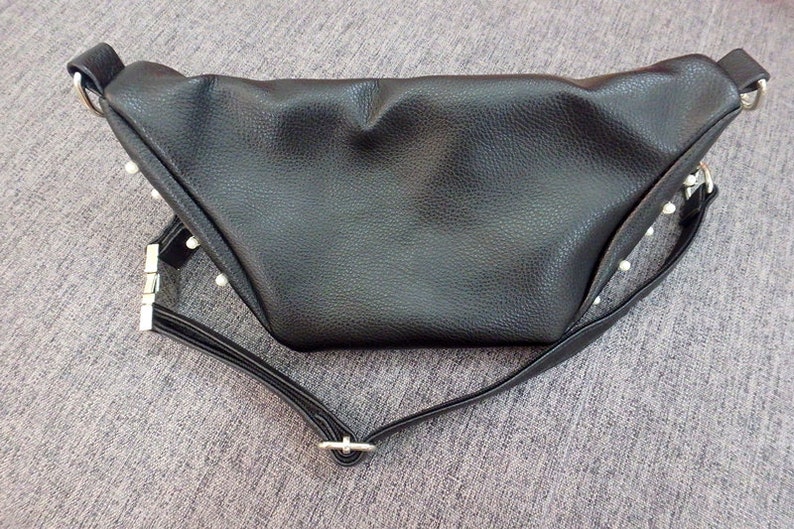 K&ouml;nnte beinhalten: Schwarze Bauchtasche mit strukturierter Oberfl&auml;che und dreieckiger Form. Die Tasche hat einen langen, verstellbaren Riemen und silberfarbene Beschl&auml;ge. Kleine, wei&szlig;e, perlenartige Verzierungen sind an den R&auml;ndern sichtbar.