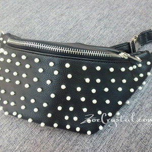 K&ouml;nnte beinhalten: Schwarze Bauchtasche mit wei&szlig;en, perlenartigen Nieten. Die Tasche hat einen silbernen Rei&szlig;verschluss und einen silbernen Kettenriemen. Die Website-Adresse "ZoeCrystal.com" ist unten rechts auf der Tasche aufgedruckt.