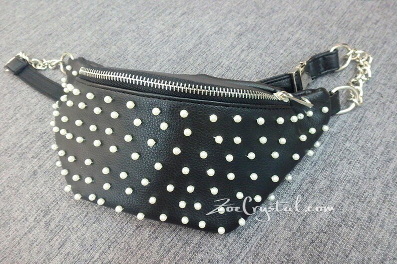 K&ouml;nnte beinhalten: Schwarze Bauchtasche mit wei&szlig;en, perlenartigen Nieten. Die Tasche hat einen silbernen Rei&szlig;verschluss und einen silbernen Kettenriemen. Die Website-Adresse "ZoeCrystal.com" ist unten rechts auf der Tasche aufgedruckt.