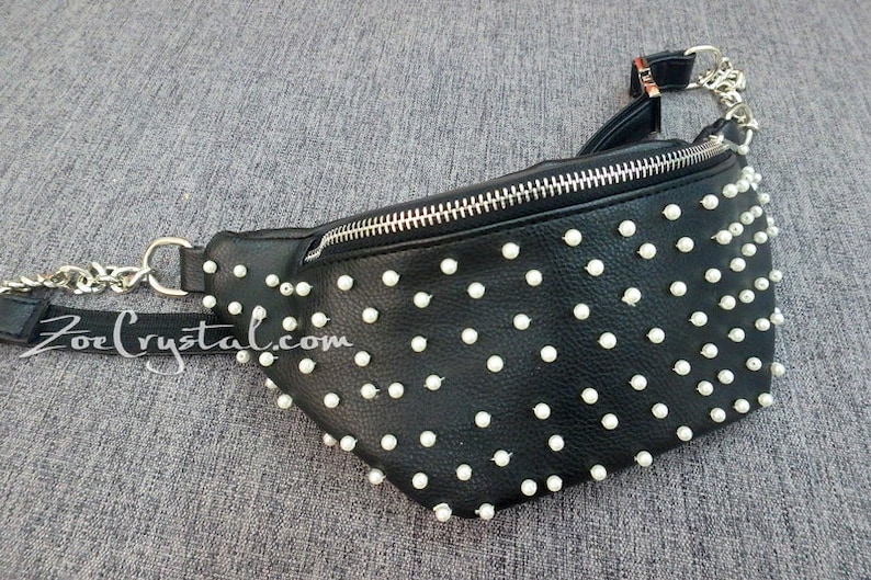 K&ouml;nnte beinhalten: Schwarze Bauchtasche mit wei&szlig;en perlenartigen Nieten und einem silbernen Kettenriemen. Die Tasche hat einen Rei&szlig;verschluss und einen schwarzen Riemen mit dem Text "ZoeCrystal.com".