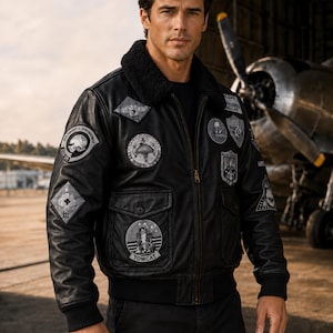 May include: A black leather bomber jacket featuring a dark faux fur collar, ribbed cuffs, and a front zip fastening. The jacket is adorned with multiple embroidered military-style patches on the chest and sleeves, including a Tomcat patch.