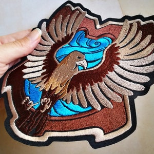 Peut inclure: Patch brodé représentant un aigle brun aux ailes déployées, un motif en spirale bleu et blanc et une bordure brune.