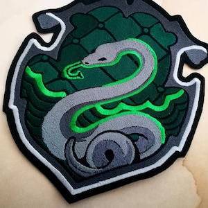 May include: Embroidered iron-on patch featuring the Hogwarts house crest for Slytherin. The crest is a green and silver shield with a silver snake coiled around it.