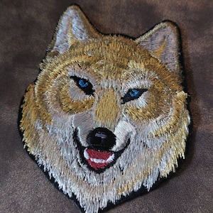 Peut inclure: Patch brodé représentant la tête d'un loup avec un motif de fourrure blanc et or et des yeux bleus. Le loup sourit avec une langue rouge.